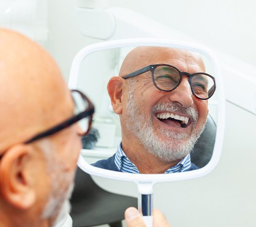 Reflection in handheld mirror of bald man with white beard in glasses in dental chair smiling